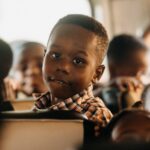 A young boy on a school bus gazes thoughtfully ahead, s